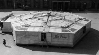Schwarz-weiß-Aufnahme eines Wanderpavillon in Form eines Hexagons mit Aufschrift &quot;Ausstellung: Die Frau&quot;. Davor eine Leiter und eine Passantin.