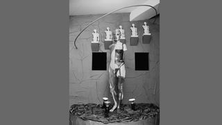 Ein weibliches Körpermodell. Die rechte Körperhälfte ist mit Reizwäsche bekleidet, die linke zeigt offen gelegte Muskelpartien. Im Hintergrund sind männliche Torsi in serieller Reihung an der Wand befestigt.