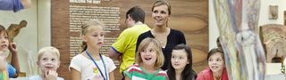 Eine Gruppe sommerlich-bunt gekleideter Kinder und ihre Betreuerin versammeln sich um die interaktive Gläsernen Frau.