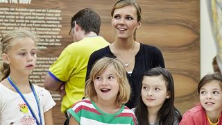 Eine Gruppe sommerlich-bunt gekleideter Kinder und ihre Betreuerin versammeln sich um die interaktive Gläsernen Frau.