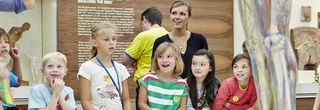 Eine Gruppe sommerlich-bunt gekleideter Kinder und ihre Betreuerin versammeln sich um die interaktive Gläsernen Frau.