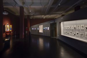 Ein dunkler Ausstellungsraum. Rechts eine lange Vitrine. Dort hängen Zeichungen zum Thema "Hass". Im Hintergrund rechts ist an einer dunkelroten Wand ein Gebetsaltar aufgebaut.  