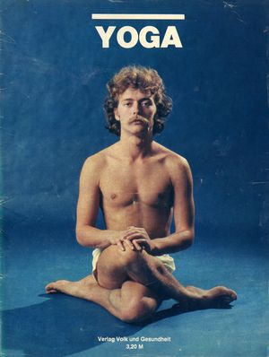 5. Raum: Ruhe Ein junger Mann mit lockigen Haaren und Schnauzbart sitzt in einer Yogapose mit überschlagenen Beinen auf dem Boden und schaut in die Kamera. Er trägt eine helle kurze Hose und ruht die Hände auf seinem Knie.