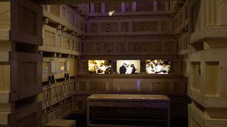 In einem Raum, dessen Wände aus hellen Holzkisten bestehen, befinden sich an der hinteren Wand drei Monitore. Auf den Monitoren laufen Filmsequenzen von Familien unterschiedlicher Nationalitäten.