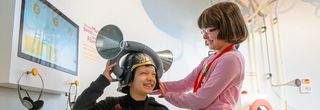 Ein Junge trägt einen Helm aus Aluminium mit zwei großen Trichtern. Neben ihm steht ein Mädchen. Beide lachen.