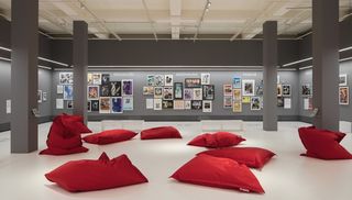 Im letzten Ausstellungsraum sind im Vordergrund rote Sitzkissen auf weißem Boden und im Hintergrund sind AIDS-Plakaten an grauen Wände.