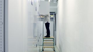 Ein Gang, rechts eine weiße Wand und links eine Reihe weißer Schränke. In der Mitte steht eine Person auf einer Leiter vor dem Schrank und ihr Oberkörper ist von einer geöffneten Schranktür verdeckt.