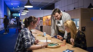 Jugendliche spielen Scrabble auf einem langen Spieltisch aus Holz in einem Raum der Spracheausstellung.