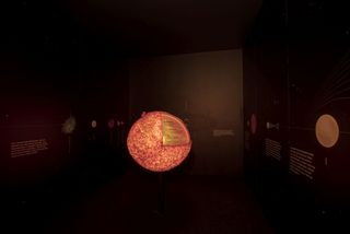 Blick in einen kleinen dunkelen Austellungsraum. In der Mitte das orange leuchtende Modell der Sonne. An den Wänden um das Sonnenmodell sind Planeten abgebildet. 