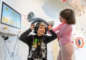 Ein Junge trägt einen Helm aus Aluminium mit zwei großen Trichtern. Neben ihm steht ein Mädchen und hilft ihm, den Helm zu halten.