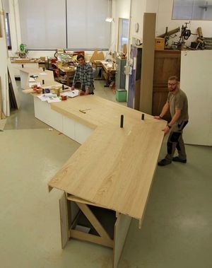 Zwei Werkstattmitarbeiter zeigen ein mäanderndes Ausstellungsmodul in Höhe eines Tisches