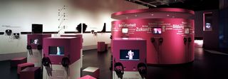 Ein dunkler Ausstellungsraum. In der Mitte es Raumes ein abgetrennter, ovaler Raum, dessen Wände pink sind. Auf den Wänden Wörter wie &quot;Zukunft&quot;, &quot;Krise&quot;, &quot;Bildung&quot;. Um den ovalen Raum stehen viereckige Säulen, auf welchen Bildschirme und Kopfhörer angebracht sind.