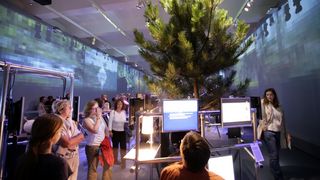 In der Mitte des Ausstellungsraumes ein circa 4 Meter hoher Nadelbaum. Der Baum ist von einem Gitter aus Stahl umgeben, an dem Monitroe befestigt sind. Sechs Besucher betrachten den Baum.