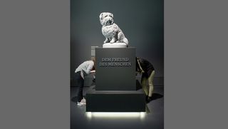 Vor einer dunkelgrauen Wand befindet sich die weiße Skulptur eines langhaarigen Hundes. Diese Skulptur steht auf einem Sockel mit der Aufschrift &quot;Dem Freund Des Menschen&quot;