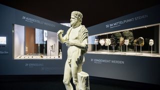 Im Hintergrund rechts steht eine Vitrine mit verschiedenen Masken. Links eine Vitrine mit verschiedenen Penisköchern. Im Vordergrund eine Statue des Anakreon mit verschnürter Vorhaut des Penis in der Ausstellung Scham.