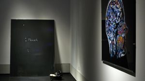 An der hinteren Wand eines Ausstellungraums eine schwarze Tafel auf der das Wort "Mensch" steht. Auf der rechten Wand ein abstraktes Gemälde, auf dem das Profil eines Menschen abgebildet ist.