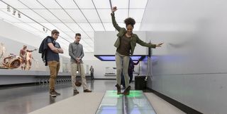 Eine junge Frau auf der metallfarbenen Balancierstrecke im Raum Bewegung, der weiß ist. Zwei junge Männer sehen ihr beim Balancieren zu.