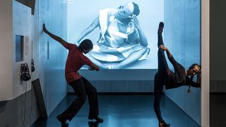 Eine junge Tänzerin und ein junger Tänzer posieren vor einer großformatigen Videoinstallation in blau und weiß in einem weißgrauen Raum.
