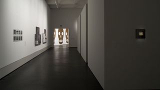 Blick in einen Ausstellungsraum. Rechts hängen Fotografien und Bilder an der weißen Wand. Im Hintergrund eine beleuchtete Wand auf der drei ca. zwei Meter hohe Totenköpfe abgebildet sind.