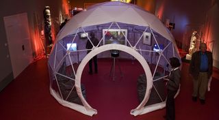 In der Mitte eines roten Ausstellungsraumes ein weißes Iglu, dessen Voderseite durchsichtig ist. In dem Iglu eine Diskokugel, zwei Sitzsäcke, zwei Monitore und zwei Bilder.