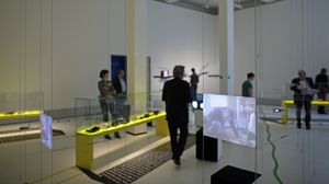 In einem weißen Ausstellungsraum hängen zwei Monitore von der Decke. In der Mitte des Raumes ein neongrüner Schaukasten. In dem Schaukasten befinden sich in einer Reihe Schuhe. 