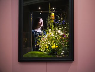 Eine Vitrine, in der ein kleiner Ausschnitt einer Wiese mit Wildblumen steht. Im Hintergrund betrachtet dies eine junge Frau.