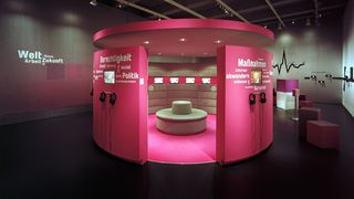 Ein dunkler Ausstellungsraum. In der Mitte es Raumes ein abgetrennter, ovaler Raum, dessen Wände pink sind. Auf den Wänden Wörter wie &quot;Gerechtigkeit&quot;, &quot;Politik&quot;, &quot;Maßnahmen&quot;. In dem ovalen Raum eine runde Sitzgelegenheit und an den Wänden Monitore.