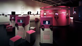 Ein dunkler Ausstellungsraum. In der Mitte es Raumes ein abgetrennter, ovaler Raum, dessen Wände pink sind. Auf den Wänden Wörter wie &quot;Zukunft&quot;, &quot;Krise&quot;, &quot;Bildung&quot;. Um den ovalen Raum stehen viereckige Säulen, auf welchen Bildschirme und Kopfhörer angebracht sind.