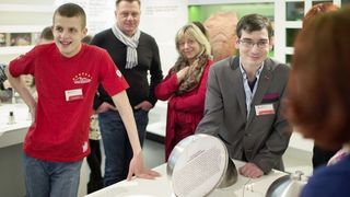 Ein junger Mann mit kurzen Haaren und ein junger Mann mit grauem Jacket bei einer Führung in Leichter Sprache. Der Mann im grauen Jacket hebt einen Servierdeckel, unter dem ein Ausstellungstext aufgedruckt ist.