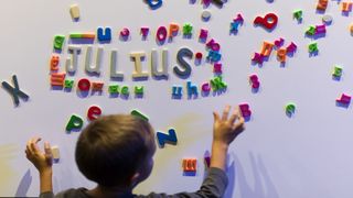 Ein Junge von hinten fotografiert setzt an einer magnetischen Buchstabenwand das Wort Julius.