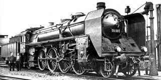 Foto: Seitenansicht einer  Schnellzuglokomotive von 1918. Links daneben stehen zwei Männer, deren Körpergröße im Vergleich zur Lokomotive ein Drittel beträgt.