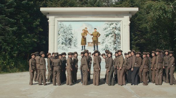 Eine große Gruppe Frauen in militärischer Uniform mit Schirmmützen steht vor einem Wandmosaik. Darauf ist eine Frau mit einem kleinen Jungen auf der Schulter zu sehen sowie ein Mann, der die Hand des Jungen hält. Alle tragen Uniformen. Hinter der Familie ist eine kleine Blockhütte sowie ein schneebedeckter, bewaldeter Berg zu sehen. Hinter dem Mosaik sieht man ebenfalls Wald.