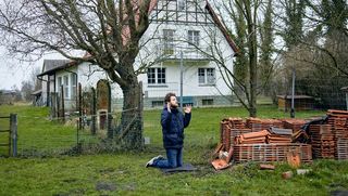 Ein Mann kniet mit gefalteten Händen in einem Garten, ein Wohnhaus und Bäume ohne Blätter im Hintergund. Rechts von ihm sind Dachziegel gestapelt.