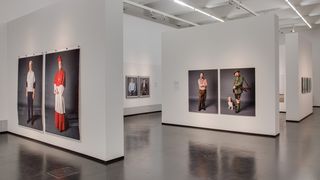Ein Ausstellungraum an dessen Wänden drei Bilder hängen, auf denen lebensgroß Menschen abgebildet sind. Auf dem vordergründigsten Bild ein Mann, der einerseits eine Jäger-Unifrom und andererseits ein Karo-Hemd und Lederhose trägt.
