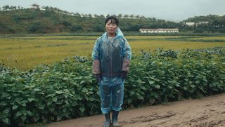 Eine Feldarbeiterin steht auf einem schlammigen Weg. Sie trägt Gummistiefel und einen transparenten blauen Plastikoverall über ihrer Kleidung. Hinter ihr ist ein Feld, einige kleine, einstöckige Gebäude und ein langgestreckter Hügel zu sehen.