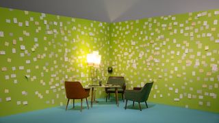 Vor einer hellgrünen Wand, an der Post-It befestigt sind, stehen drei dunkelgrüne Sessel, ein Tisch und eine Stehlampe.