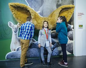 Ein Mädchen steht in der Mitte und hat überdimensionale Ohren aus Fell aufgesetzt. Rechts und links sprechen zwei andere Kinder in die Ohren. An dieser Station wird erfahrbar gemacht, wie man mit großen Ohren hört.