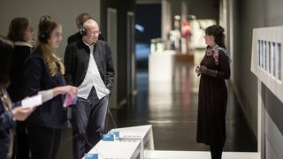 In einem Ausstellungsraum steht recht eine Frau mit lockigen Haaren vor einer Wand mit Bildern und erzählt einer Gruppe von Personen, die links im Bild sind, etwas. Im Hintergrund ist ein weiterer Raum zu erkennen.