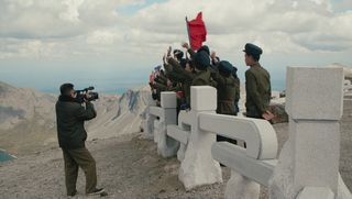 Eine Gruppe Koreaner:innen steht einer Balustrade auf einem Bergplateau. Sie tragen militärische Kleidung. Sie winken und schwenken eine große rote Fahne. Falst alle halten ein kleines rotes Buch, etwa im Passformat. Sie werden von einem Kameramann gefilmt. Im Hintergrund ist ein Bergkessel unter einem bewölkten Himmel zu sehen.