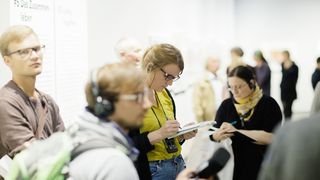 Im Zentrum eine junge Frau mit Kopfhörern, die sich Notizen macht. Vor und hinter ihr sind weitere Beucher.