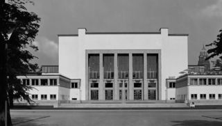 Schwarzweißes Foto im Hintergrund mit dem Museum von vorn. Im Vordergrund stehen links und rechts Bäume.