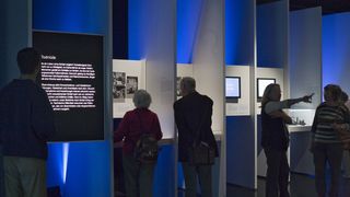 Links betrachtet ein Mann eine Informationstafel mit der Überschrift &quot;Todmüde&quot;. In der Mitte betrachten eine ältere Dame und ein älterer Herr Fotografien an der Wand. Rechts weitere Bescher. 